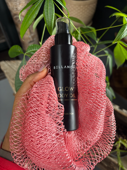 Bellangi Glow Body Oil bottle held in a pink mesh bag with plants in the background